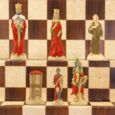 The Red side of a handpainted chess set featuring characters from the Arthurian legend, placed on a checkered board. The set includes figures representing King Arthur, Queen Guinevere, Merlin the Magician, Sir Lancelot the Knight, & the Castle of Camelot as the rook.