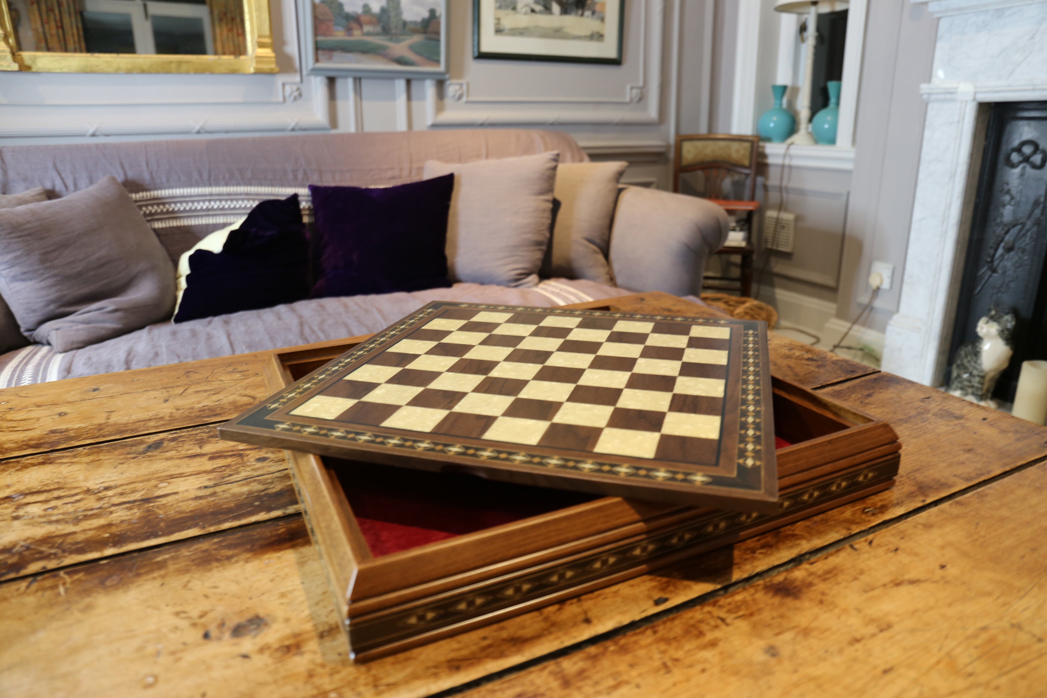 A polished wooden chess board with a checkered pattern, featuring eco mother of pearl details on the edges which contains storage for chess pieces below. This is on a wooden table in a rustic abode.