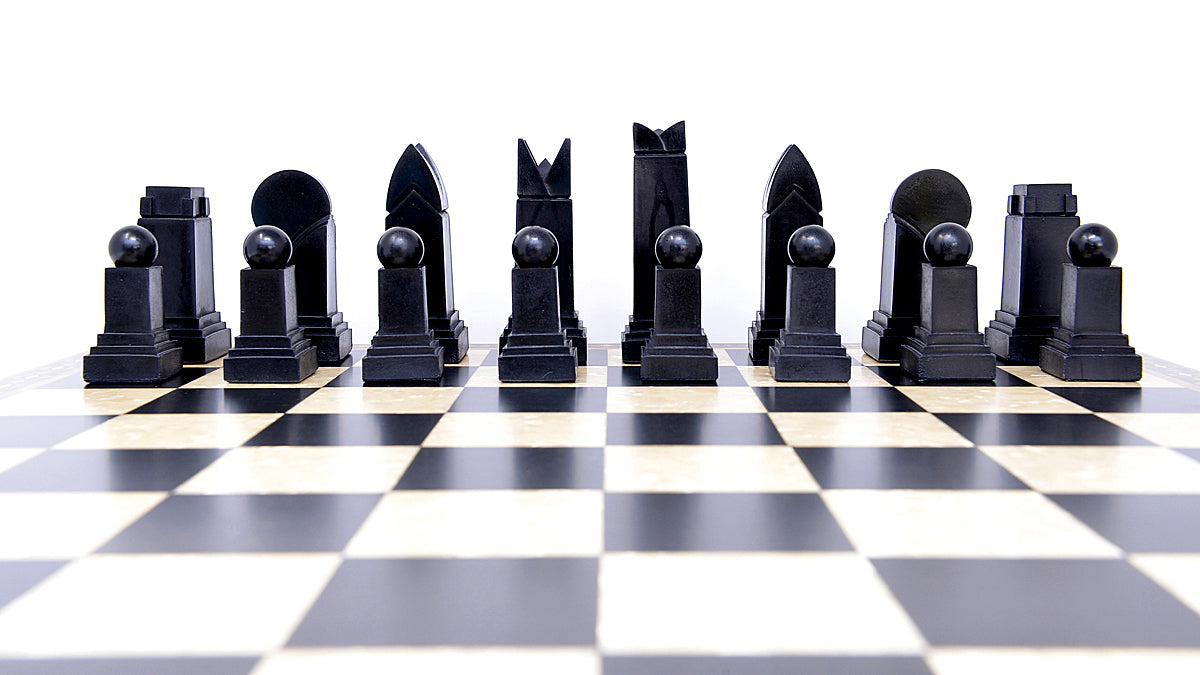 The black side of a handmade chess set displayed on a checkered board. The set includes various shapes of pieces, such as rooks, knights, bishops, and pawns, showcasing an Art Deco design style.