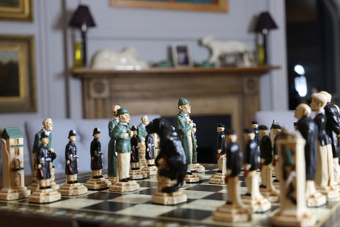 Hand painted chess set featuring characters resembling Sherlock Holmes and other figures, placed on a wooden chessboard with a manor backdrop.