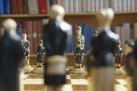 Hand painted chess set featuring characters resembling Sherlock Holmes and other figures, placed on a wooden chessboard. With a bookshelf in the background.
